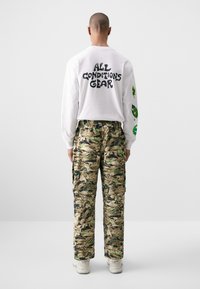 Hvit langermet skjorte med svart tekst "ALL CONDITIONS GEAR" på ryggen; cargo-bukser med kamuflasjemønster og grønne detaljer.