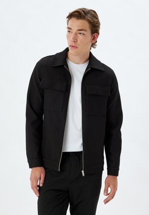 Chaqueta negra de canalé con cierre de cremallera, dos bolsillos en el pecho y un cuello plano. Lleva puesta sobre una camiseta blanca y combinada con pantalones negros.