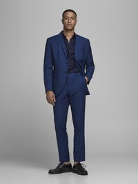 Navy blue suit made of textured fabric, featuring a single-breasted jacket and slim-fit trousers, paired with a dark shirt and black shoes.