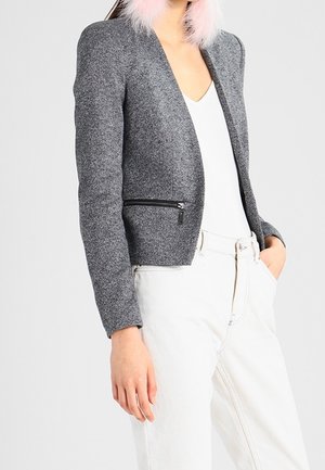 Femme portant une veste grise texturée avec une poche zippée noire, un haut blanc, un pantalon blanc, et des boucles d'oreilles ou un accessoire de col rose duveteux.