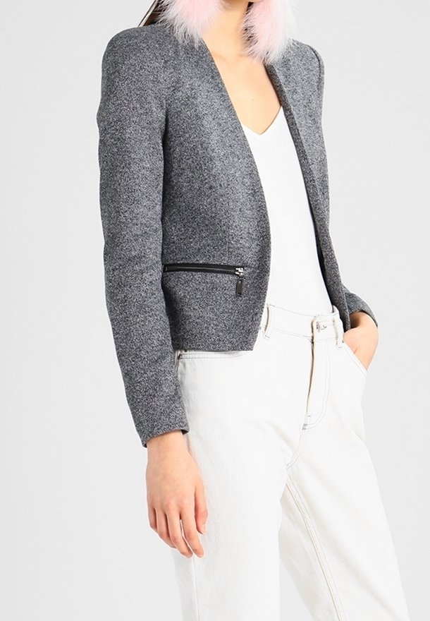 Femme portant une veste grise texturée avec une poche zippée noire, un haut blanc, un pantalon blanc, et des boucles d'oreilles ou un accessoire de col rose duveteux.