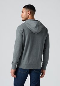 Uomo visto di spalle che indossa una felpa grigia con cappuccio e jeans blu su uno sfondo grigio chiaro uniforme.