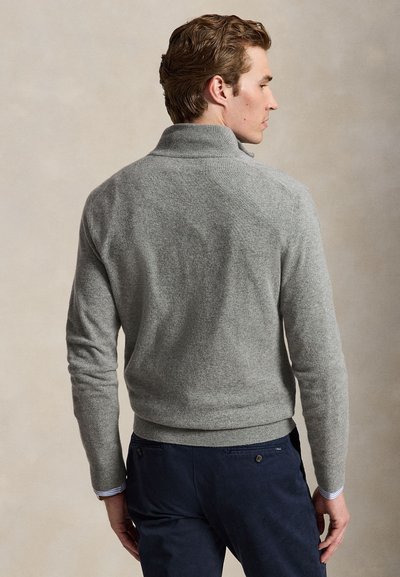 Hombre con cabello corto y castaño lleva un suéter gris y pantalones azul oscuro, de pie de perfil con un fondo beige neutro.