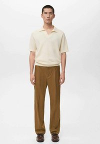 Polo crème à manches courtes avec col en V et tissu texturé, associé à un pantalon marron à jambes droites et des chaussures foncées à lacets.
