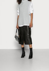 Pull à col roulé gris côtelé avec détail de boutons, superposé à une chemise blanche, associé à une jupe midi en cuir noire et des bottines noires.