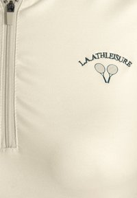Heller beiger Reißverschlussoberteil aus glattem Stoff. Mit gesticktem Text "LAATHLEISURE" und zwei gekreuzten Tennisschlägern darunter.