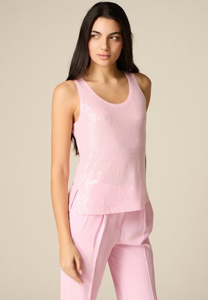 Kvinde med langt, mørkt hår iført en ærmeløs, skinnende lyserød top og matchende lyserøde bukser med høj talje, stående foran en beige baggrund.