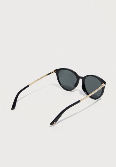 Μαύρα γυαλιά ηλίου σε σχήμα γάτας με σκούρους φακούς και λεπτούς χρυσούς βραχίονες που ακουμπούν σε λευκή επιφάνεια.