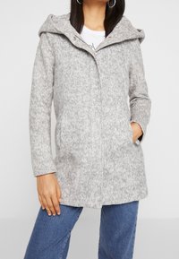 Femme portant un manteau en laine à capuche gris clair avec des poches sur le devant et un jean bleu, une main dans la poche et l'autre détendue.