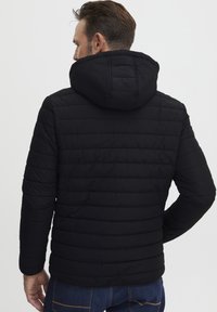 Črna napihnjena jakna s kapuco, ki ima prešito zasnovo, rebraste manšete in sproščen kroj. Zadnji pogled prikazuje vodoravne šive.