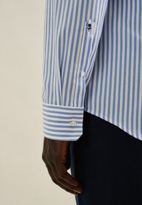 Camicia a righe azzurro chiaro e bianca con polsini abbottonati. Presenta un emblema floreale nero sulla manica. Tessuto in cotone liscio.