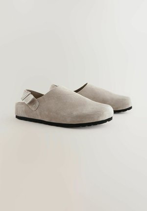 Suede clogs in light gray with a smooth texture, rounded toe, adjustable ankle strap, and black rubber sole.