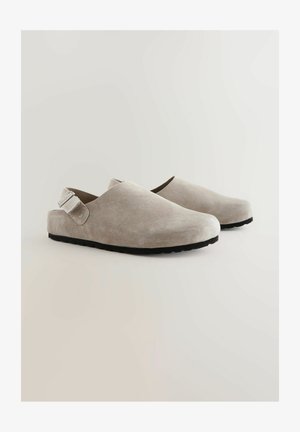 Saboti din suede de culoare gri deschis, cu o textură netedă, vârf rotund, curea de gleznă ajustabilă și talpă din cauciuc negru.