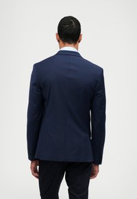 Tamsiai mėlynas pritaikytas kostiumo švarkas su tvirtu siluetu, vienu skilimu nugaroje ir sagstomais rankogaliais. Lygus audinio paviršius.