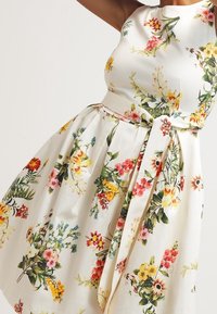 Vestido con estampado floral que presenta un corpiño ajustado, falda acampanada y un cinturón de tela. Los colores incluyen blanco, amarillo, rosa y verde.