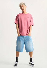 Camiseta rosa de algodón con mangas cortas, combinada con pantalones cortos de mezclilla azul claro. Zapatillas negras y blancas con calcetines negros hasta la mitad de la pantorrilla.