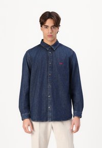 Dark denim button-up shirt with a collar, long sleeves, and a small red logo detail on the chest. Structured fit with a straight hem.