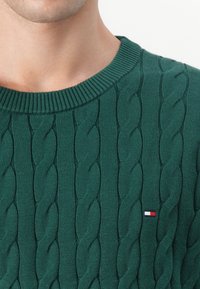 Zeleno pleteno pulover z vrvnim vzorcem, rebrastim ovratnikom in majhnim logotipom v rdeči, beli in modri barvi. Mehka tekstura in prilega forma.