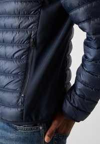 Marineblå pufferjakke med horisontalt quiltet design, sidelomme med glidelås, glatt tekstur og en tettsittende silhuett.