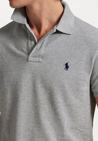 Camisa polo gris hecha de tejido texturizado, con un cuello clásico, dos botones y un logo bordado en azul marino en el pecho.