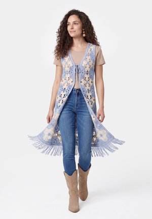 Kvinde iført blå og cremefarvet hæklevest med frynser over beige top, blå jeans og brune cowboystøvler går fremad.