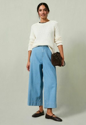 Heller cremefarbener Pullover mit strukturiertem Strickmuster, kombiniert mit blauen, gestreiften Weitbein-Hosen und dunkelbrauner Schultertasche. Schlichte schwarze Ballerinas.