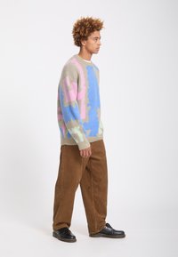 Jeune homme aux cheveux bouclés porte un pull coloré à motifs, un pantalon en velours marron et des chaussures noires, debout de profil sur un fond blanc.
