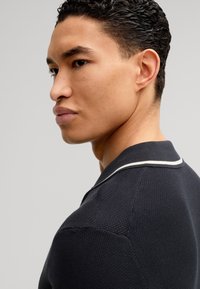 Polo shirt navy con tessuto texturizzato, con colletto rifinito in bianco. Focus sui dettagli della spalla e del collo, mostrato di lato.