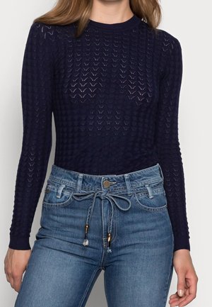 Femme portant un haut à manches longues texturé bleu marine associé à un jean bleu taille haute noué avec une ceinture à cordon en denim.