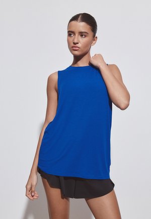 Ung kvinde i ærmeløs kornblå top og sorte shorts, stående med en hånd nær skulderen, kiggende til siden mod en ensfarvet baggrund.