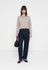 Cárdigan corto beige con botones en la parte delantera y bordados; combinado con jeans de pierna ancha azul oscuro. La modelo sostiene un pequeño bolso negro y lleva puestos unos zapatos burdeos.