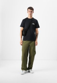 Un jeune homme se tient devant un fond sobre, portant un t-shirt noir, un pantalon cargo vert et des baskets blanches, avec un petit sac noir en bandoulière.