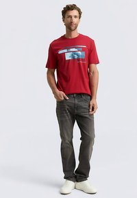 Červené bavlněné tričko s grafickou potiskem, doplněné tmavě šedými džíny a bílými teniskami. Střih je volný a pohodlný, s krátkými rukávy.