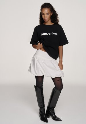 Kobieta o kręconych włosach, ubrana w czarną koszulkę z napisem „GIRL'S GIRL”, białą bufiastą spódnicę, czarne rajstopy i czarne kozaki sięgające do kolan, stojąca pewnie.