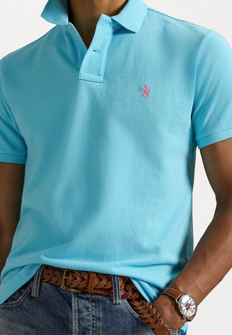 Hellblaues Poloshirt mit strukturierter Oberfläche und pinkem gesticktem Logo. Getragen mit einem braunen geflochtenen Gürtel und Denim-Shorts. Silberne Uhr am Handgelenk.