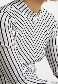 Långärmad topp i vitt och svarta ränder, med hög krage och chevronmönster, tillverkad av ett strukturerat tyg.