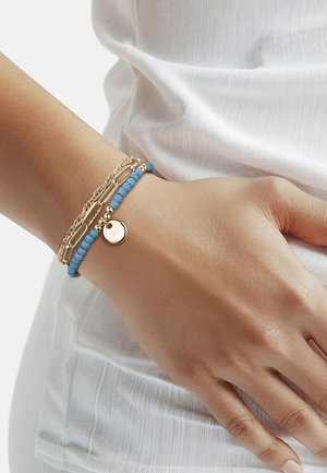 Poignet avec deux bracelets : une chaîne en or et un bracelet bleu à perles avec un pendentif rond en or, sur un tissu blanc texturé.