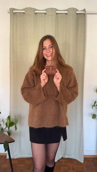 Brown fluffy sweater, black skirt, holding a small brown textured wallet with a zipper. Background features a light curtain and plants.