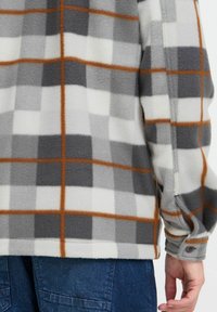 Camiseta de forro polar con un patrón de cuadros en gris, blanco y naranja; presenta puños abotonados y laterales abiertos; textura suave y corte relajado.