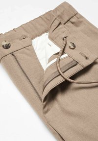 Pantalon beige avec une ceinture élastique, un cordon, une fermeture par bouton, une braguette zippée ouverte et une doublure de poche blanche visible.