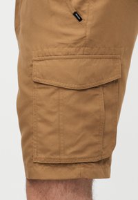 Μπεζ cargo σορτς με μεγάλη τσέπη με καπάκι στο πλάι και μικρή μαύρη ετικέτα "Rip Curl" πάνω από την τσέπη.