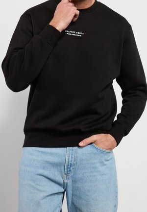 Mann trägt einen schwarzen Kapuzenpulli mit dem Text "Crafted Goods Pier One Denim", helle blaue Jeans, eine Hand in der Tasche, die andere berührt das Kinn.