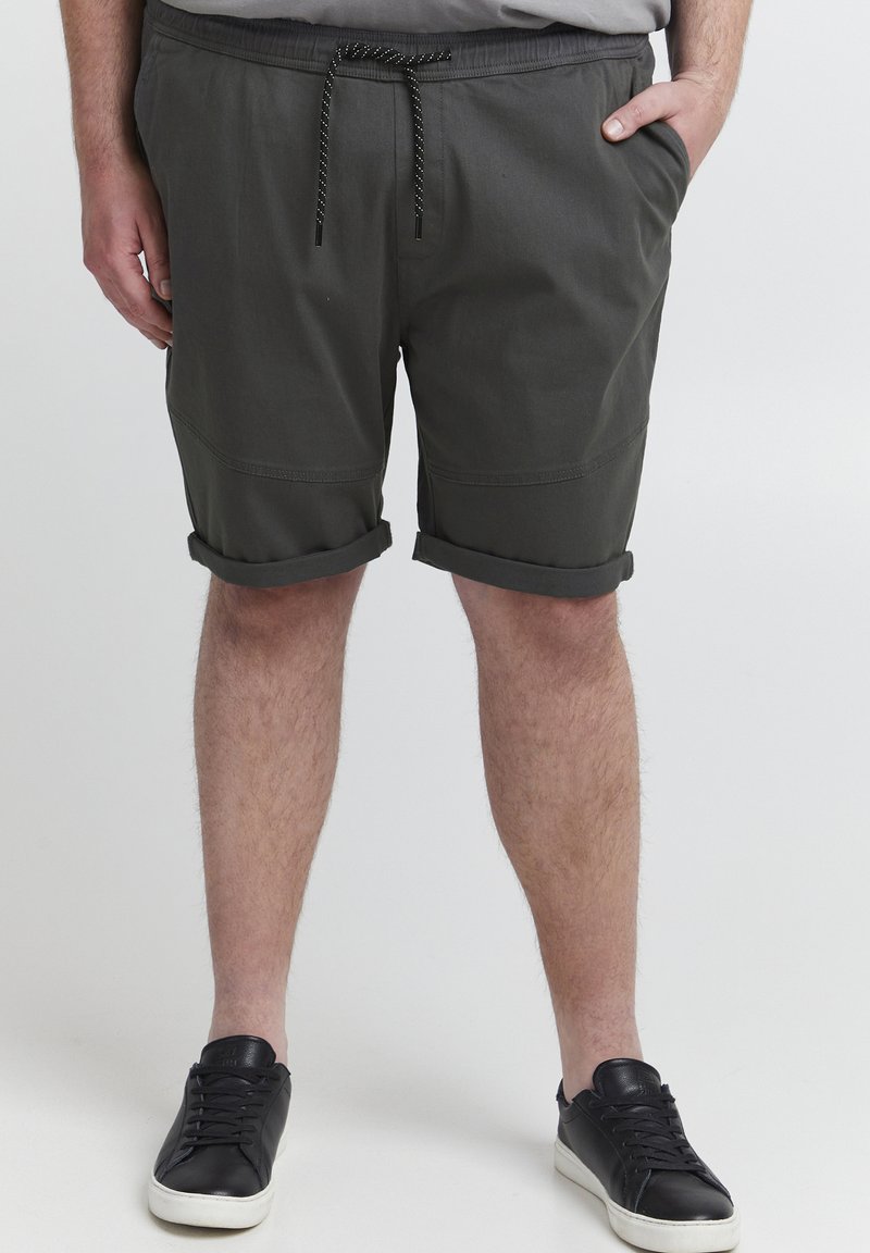 Dunkelgrüne Shorts mit umgeschlagenen Säumen, elastischem Bund und Kordelzug mit schwarz-weißen Akzenten. Getragen mit schwarzen Turnschuhen.