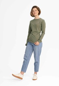 Langärmliges olivgrünes T-Shirt aus Baumwolle, kombiniert mit blauen Jeans und hellen Sneakers; das Modell steht in einer entspannten Pose.