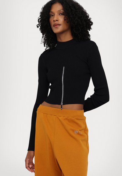 Fekete bordázott cropped top magas nyakkivágással és cipzár részlettel; narancssárga melegítőnadrággal párosítva, amelyen egy kis hímzett minta található.
