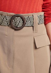 Woven black and cream belt with a circular brown buckle over tan fabric trousers featuring a side pocket and textured red ribbed top.