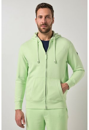 Mand iført lysegrøn hættejakke med lynlås og matchende bukser, stående med den ene hånd i lommen mod en ensfarvet baggrund.