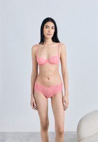 Rožinės nėriniuotos liemenėlės ir kelnaičių rinkinys su geometriniu raštu, banguotais kraštais ir reguliuojamais petnešėlėmis, fone yra neutrali spalva.