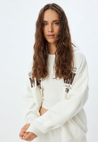 Weißer, kurzer Hoodie mit lockerer Passform, der braunen grafischen Text aufweist. Das Material wirkt weich und strukturiert und ergänzt ein schlichtes Design.