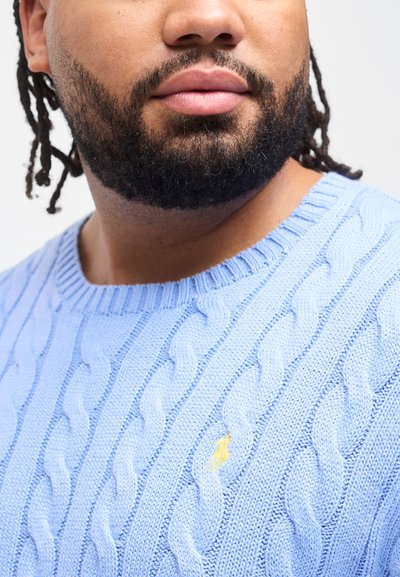 Κοντινή λήψη ενός άνδρα με γενειάδα και dreadlocks, που φοράει ένα ανοιχτό μπλε πουλόβερ με πλέξη καλώδιο και μικρό κίτρινο κεντημένο λογότυπο.